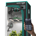 Indoor grow tent with ventilation fan, LED light, circulation fan, plants, and a smartphone showing control settings